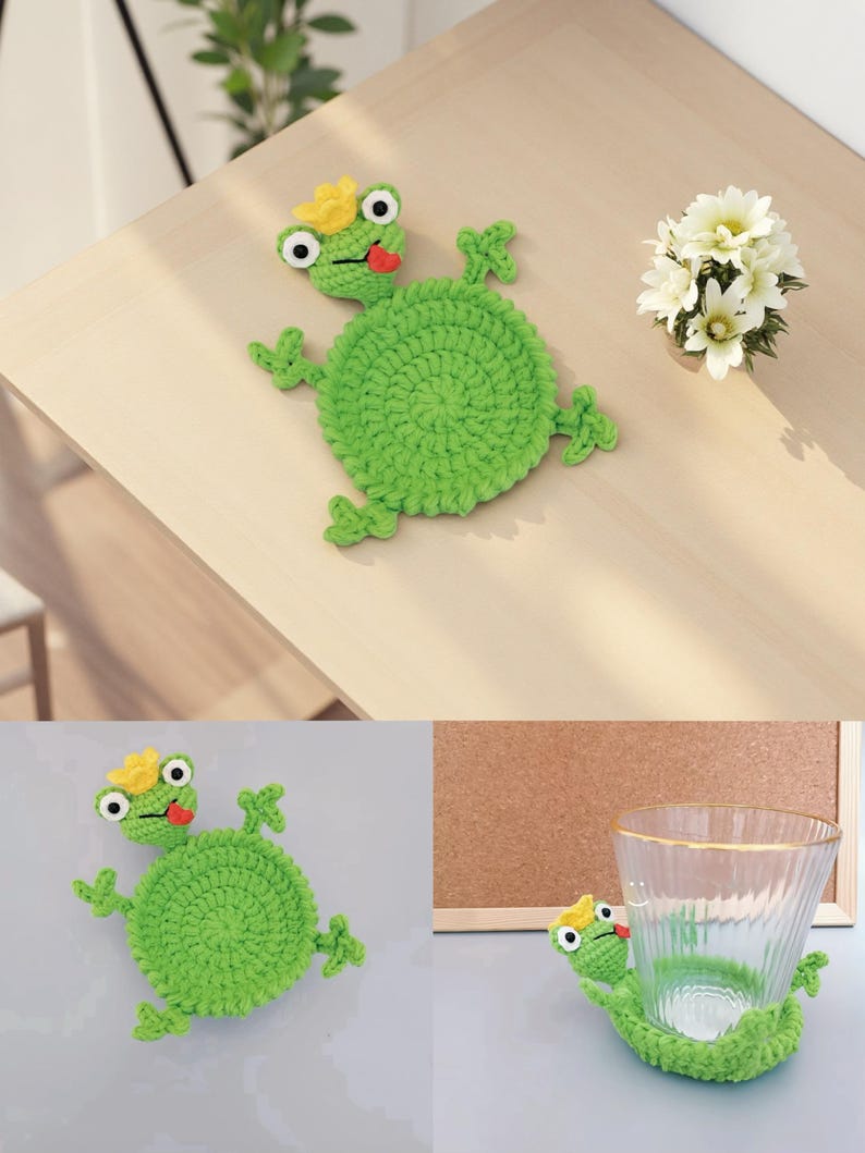 May include: A green crocheted frog coaster with a yellow crown and red tongue. The frog has large white-rimmed eyes and small green limbs. The coaster is round and sits on a light-colored wooden surface. A small bouquet of white flowers is also present.