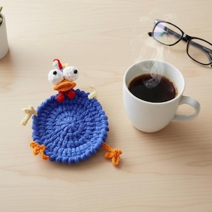 Könnte beinhalten: Ein blauer, gehäkelter Hühneruntersetzer mit weißen Augen, einem orangefarbenen Schnabel und orangefarbenen Füßen. Eine weiße Tasse Kaffee dampft neben dem Untersetzer. Eine schwarze Brille liegt auf der Holzoberfläche.