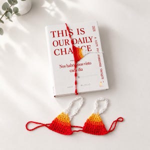 Könnte beinhalten: Ein weißes Buch mit dem Titel THIS IS OUR DAILY CHANCE in roter Schrift, umwickelt mit einem gehäkelten Lesezeichen in Rot, Orange und Weiß. Unter dem Buch liegt ein passendes gehäkeltes Bikini-Oberteil in Rot, Orange und Weiß mit weißen Trägern.