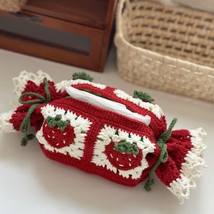 Puede incluir: Una funda de caja de pañuelos de ganchillo roja y blanca hecha a mano con forma de envoltorio de caramelos. La funda presenta diseños de fresas y detalles en verde. La funda de la caja de pañuelos está atada con hilo verde.