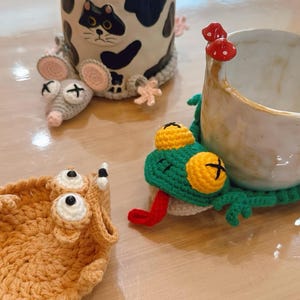 May include: A ceramic mug with a black and white cow pattern and a cat face design. The mug is accompanied by crocheted cup cozies in the shape of a mouse, a frog, and a creature with large eyes. The frog has a red tongue.