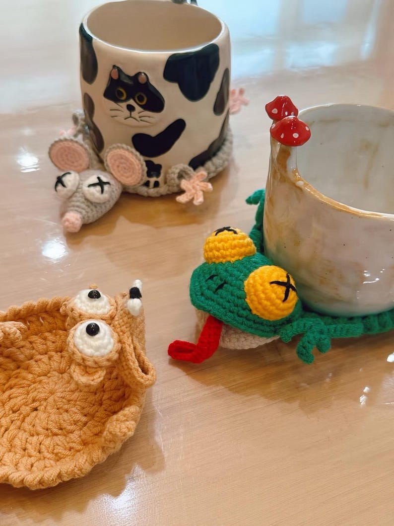 May include: A ceramic mug with a black and white cow pattern and a cat face design. The mug is accompanied by crocheted cup cozies in the shape of a mouse, a frog, and a creature with large eyes. The frog has a red tongue.
