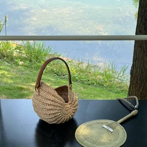May include: A woven, beige seashell-shaped handbag with a brown leather handle sits on a black surface next to a matching hand fan. The fan has a woven circular design and a long handle. The background shows a body of water and greenery.