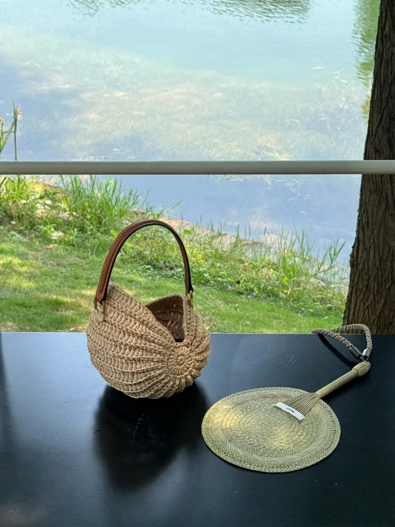 May include: A woven, beige seashell-shaped handbag with a brown leather handle sits on a black surface next to a matching hand fan. The fan has a woven circular design and a long handle. The background shows a body of water and greenery.