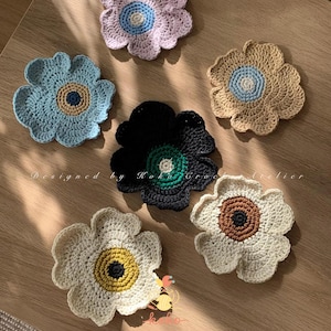 Könnte beinhalten: Eine Sammlung handgefertigter gehäkelter Blumenuntersetzer in verschiedenen Farben, darunter Blau, Schwarz, Beige und Weiß. Jeder Untersetzer hat eine kontrastierende Mitte und ein Blütenblattdesign. Der Text "Designed by Koko Crochet Atelier" ist sichtbar.