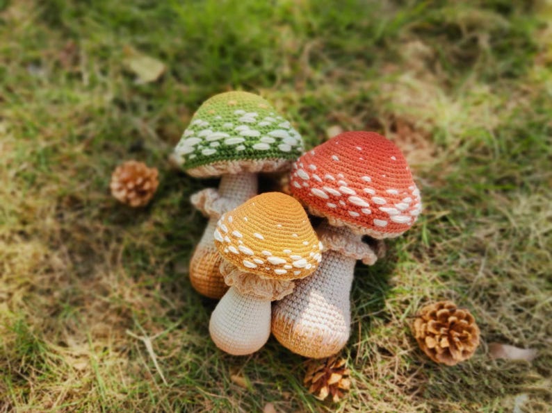 Puede incluir: Un grupo de tres setas de ganchillo con sombreros salpicados de blanco en tonos verdes, naranjas y rojos. Las setas est&aacute;n colocadas sobre hierba verde, con pi&ntilde;as cerca. Las setas hechas a mano tienen tallos blancos.