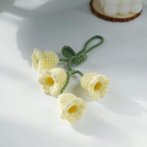 Könnte beinhalten: Handgefertigtes gehäkeltes Blumenarrangement mit vier gelben, glockenförmigen Blüten mit grünen Stielen und Blättern. Die Blumen haben ein strukturiertes Aussehen, vor einem weißen Hintergrund.