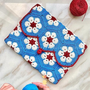 Peut inclure: Une pochette au crochet faite à la main avec un motif floral bleu, blanc et rouge. La pochette a une fermeture à rabat avec un bouton et une bordure rouges. Une pelote de fil rouge est visible en arrière-plan.