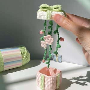 Może przedstawiać: Ręcznie robiona, szydełkowa donica wisząca w kształcie jasnoróżowego kwadratu z zielonymi akcentami. Donica zawieszona jest na białym wierzchu z zieloną kokardą i posiada zielone pnącza z różowymi kwiatami i niebieskim motylem.