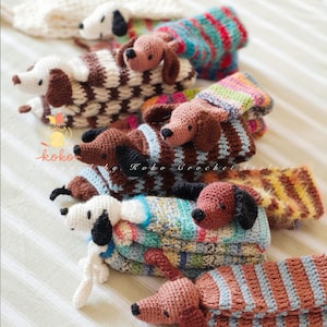 May include: A collection of handmade crochet dachshund dogs in various colors and patterns. The dogs have brown, black, and white bodies with matching ears and noses. Some dogs wear striped or patterned sweaters. The text "koko" is visible.