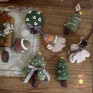 May include: A collection of handmade crochet ornaments, including green Christmas trees, birds, and a tree with white flowers. The ornaments are in various colors and designs, with brown tree trunks and decorative bows. The image includes the text "Designed by Koko Crochet."