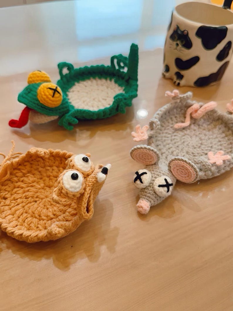 May include: A collection of handmade crocheted coasters in the shape of animals. Features a green frog, a tan creature, and a grey mouse. A cat-themed mug with black and white spots is also visible in the image.