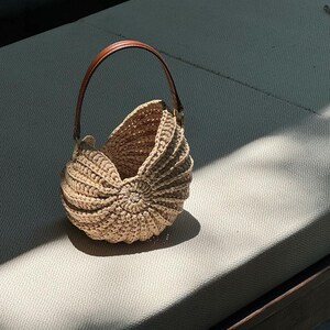May include: A beige crochet handbag with a brown leather handle. The bag has a unique shell-like design with a circular pattern at the base. The handle is arched and attached to the bag with gold-colored hardware. The bag is sitting on a light gray surface.