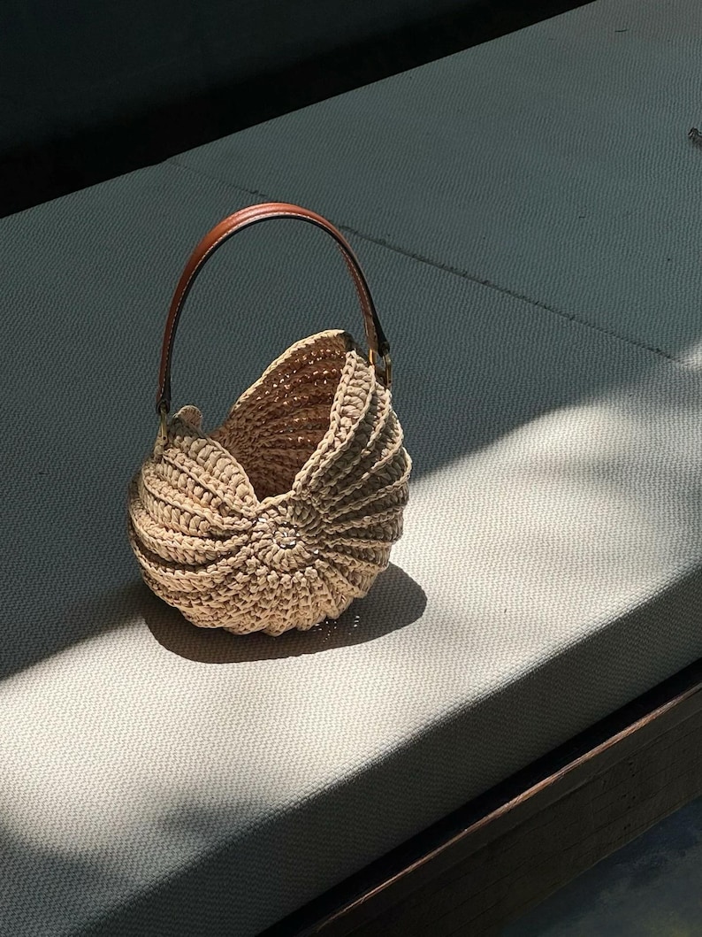 May include: A beige crochet handbag with a brown leather handle. The bag has a unique shell-like design with a circular pattern at the base. The handle is arched and attached to the bag with gold-colored hardware. The bag is sitting on a light gray surface.
