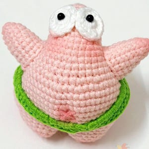 Op de afbeelding: Een roze gehaakt zeesterfiguur met twee grote witte ogen met zwarte pupillen. Het figuur heeft twee uitgestrekte armen en draagt een felgroene gehaakte tailleband rond het midden. De textuur is uniform en gestikt.