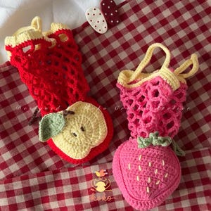 May include: Crocheted fruit-shaped bags, one red apple and one pink strawberry, with yellow accents and green leaves. The apple bag has a brown stem. The strawberry bag has a handle. The bags are on a red and white checkered surface.