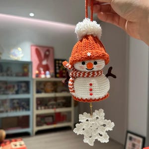 May include: A handmade crochet snowman ornament. The snowman wears an orange hat with a white pom-pom, a scarf with brown and white stripes, and a white body with red buttons. A white snowflake hangs below.