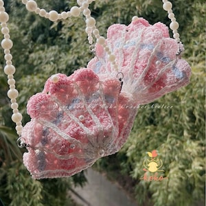 May include: A crochet seashell-shaped purse in pink, white, and blue, decorated with faux pearls. It features a pearl chain strap and the text "Designed by Koko Crochet Atelier". The purse is handmade.