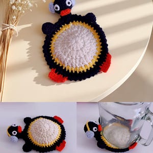 May include: A handmade crochet coaster in the shape of a penguin. The coaster features a white center, a yellow and black border, and red feet. The penguin has a black head with white eyes and blue earmuffs.