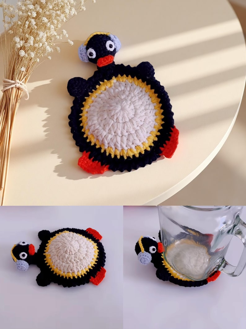 May include: A handmade crochet coaster in the shape of a penguin. The coaster features a white center, a yellow and black border, and red feet. The penguin has a black head with white eyes and blue earmuffs.