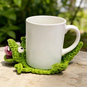 Könnte beinhalten: Eine weiße Keramik-Tasse steht auf einem handgefertigten grünen Krokodil-Untersetzer aus Häkelarbeit. Der Untersetzer hat weiße Augen und eine rosa Zunge, was einen skurrilen Touch verleiht. Die Tasse hat ein schlichtes, klassisches Design.