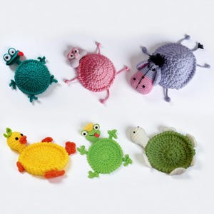 May include: Six handmade crochet animal coasters are displayed. The coasters feature a green frog, a pink pig, a purple donkey, a yellow duck, a green frog, and a green turtle. Each coaster has a circular body with animal features.