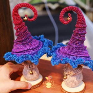 May include: Two handmade crochet mushroom ornaments. The caps are a red to purple gradient, with blue frilled edges. Each has a small gold bell and sits on a wooden base. The stems are beige with a bell.