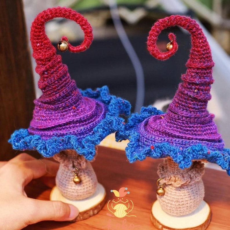 May include: Two whimsical, handmade crochet mushroom figures. The mushroom caps are a gradient of red to purple, with blue ruffled edges. Each has a small gold bell and sits on a wooden base. The stems are beige with a bell.
