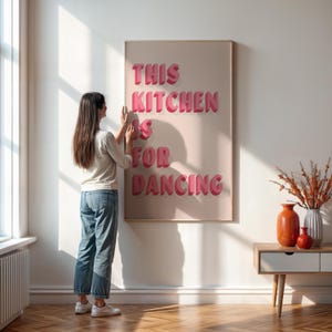 May include: A framed wall art piece with the text "THIS KITCHEN IS FOR DANCING" in pink 3D lettering. The artwork is displayed on a light pink background. A person is seen adjusting the artwork on a white wall.