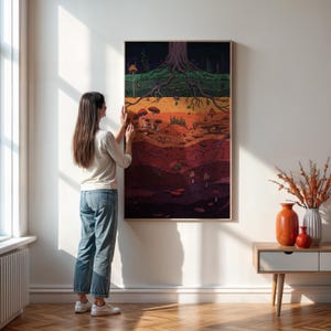 May include: A framed art print depicting a cross-section of earth with a tree and mushrooms. The artwork features a color palette of browns, oranges, and greens. The print is displayed on a white wall, with a woman adjusting the frame.