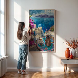 May include: A framed art print depicting a coastal scene with white buildings, blue domes, and a turquoise sea. A copper coffee pot and a cup of coffee sit on a table in the foreground. The print is being hung on a white wall.