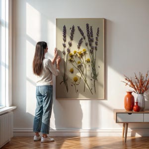 May include: A framed botanical print featuring lavender and yellow chamomile flowers. The artwork is displayed on a light-colored wall, with a person adjusting the frame. The room is illuminated by natural light, with a wooden side table and vases in the background.