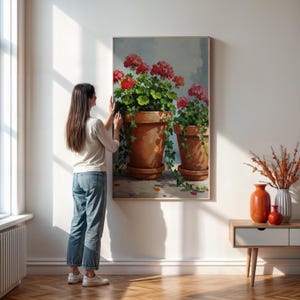 May include: A framed painting depicts two terracotta pots overflowing with vibrant red geraniums and lush green foliage. The artwork, with a light-colored background, is displayed on a white wall, adding a touch of nature-inspired decor to the space.