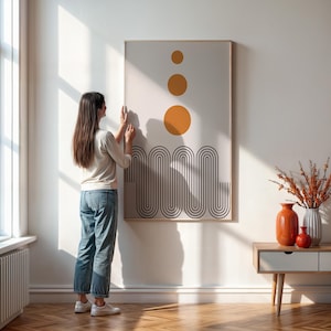 May include: A person hangs a framed abstract print on a white wall. The print features a series of black curved lines and three orange circles.