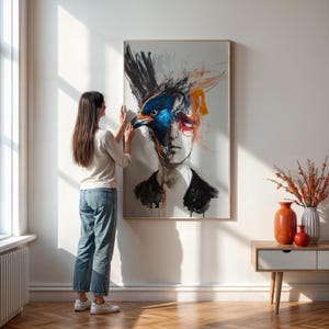 May include: A large framed artwork featuring a surreal portrait of a person with a bird's head. The artwork uses a mix of blue, black, orange, and white colors. The person is wearing a black jacket. The artwork is hanging on a white wall.