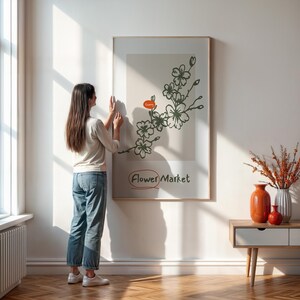 May include: A woman hangs a framed print on a white wall. The print features a black and white illustration of a flowering branch with the text "Flower Market" in a speech bubble.