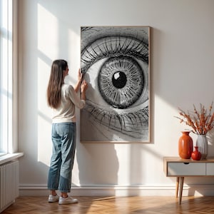 May include: A large, framed black and white print of a human eye. The artwork is being hung on a white wall. The eye is highly detailed, with visible eyelashes and iris. The frame is a light wood color.