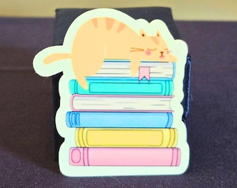 May include: A sticker of an orange cat sleeping on top of a stack of colorful books. The cat has a pink bookmark in its mouth.