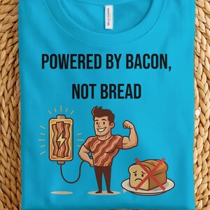 May include: A turquoise t-shirt with the text "POWERED BY BACON, NOT BREAD" and a cartoon illustration of a flexing man with bacon on his torso. The shirt also features a bacon-shaped battery and a piece of bread with a red "no" symbol.