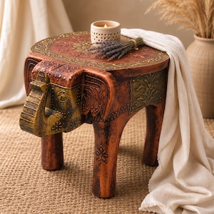 Peut inclure: Un tabouret en bois marron en forme d'éléphant avec des accents peints en doré sur la tête et la trompe. Le dessus circulaire présente des sculptures florales complexes et un motif de bordure. Le tabouret repose sur quatre pieds robustes.