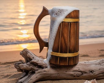 Può includere: Un boccale di birra in legno con finitura marrone scuro e manico decorativo. Il boccale è pieno di birra schiumosa, che trabocca sul lato. Il boccale è appoggiato su legno trasportato sulla spiaggia sabbiosa con l'oceano e il tramonto sullo sfondo.