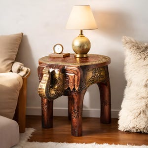 Puede incluir: Una mesa auxiliar de madera artesanal con diseño de elefante. La mesa es marrón con detalles dorados y tiene una tapa redonda. Una lámpara dorada y un pequeño reloj están encima. Una pieza de mobiliario única.