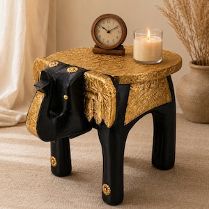 May include: A black and gold elephant-shaped side table with a round top featuring an intricate embossed pattern. The table has four sturdy legs, with gold floral accents on the ears, trunk, and feet. A wooden analogue clock and a lit candle sit on top.