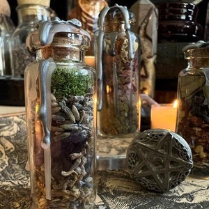 Op de afbeelding: Drie glazen flessen gevuld met gedroogde kruiden en bloemen, verzegeld met was. De flessen staan op een tafel met een pentagramvormige metalen hanger op de voorgrond.