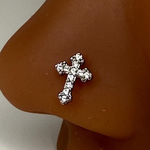 May include: A silver-colored nose stud in the shape of a cross, encrusted with small, clear gemstones. The cross is positioned on the side of a brown nose, with a white background.