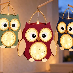 May include: Three owl-shaped lanterns in green, maroon, and blue. Each lantern features large, illuminated circular eyes and a belly with gold glitter accents, a small brown beak, and light-coloured wings and feet, suspended by a gold rope handle.
