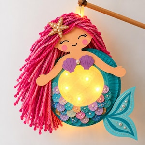 May include: A handcrafted mermaid lantern featuring long, bright pink yarn hair adorned with a gold starfish hair clip. The mermaid has a light-up yellow circular body, purple shell top, and a teal paper tail decorated with glittery scales and pearls.
