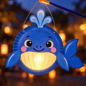 May include: A blue whale-shaped paper lantern featuring a circular body with a light-up yellow belly, pink cheeks, and a water spout on top. The lantern is attached to a thin wire hanger and a blue stick, displaying a cartoon-style whale face.