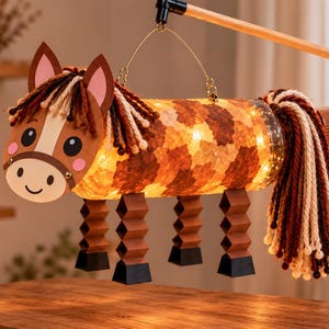 Puede incluir: Un farol con forma de caballo hecho con un cuerpo cilíndrico y una luz cálida en su interior. El caballo tiene una cabeza de papel marrón con una mancha blanca, mejillas rosadas y crines de lana. Posee cuatro patas plegadas en acordeón.