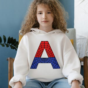 May include: A white hoodie with a red and blue spiderweb letter A on the front. The hoodie is worn by a young person with curly hair sitting on a wooden chair.